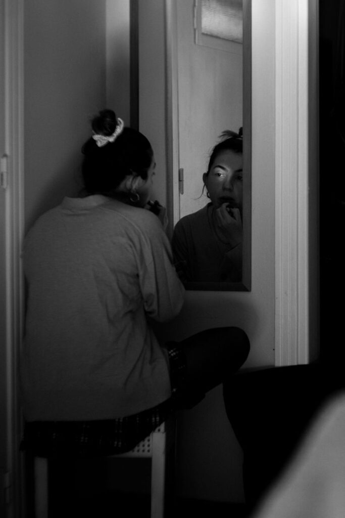A woman sits quietly in front of a mirror, applying makeup in a dimly lit room, her reflection suggesting introspection and emotional restraint.