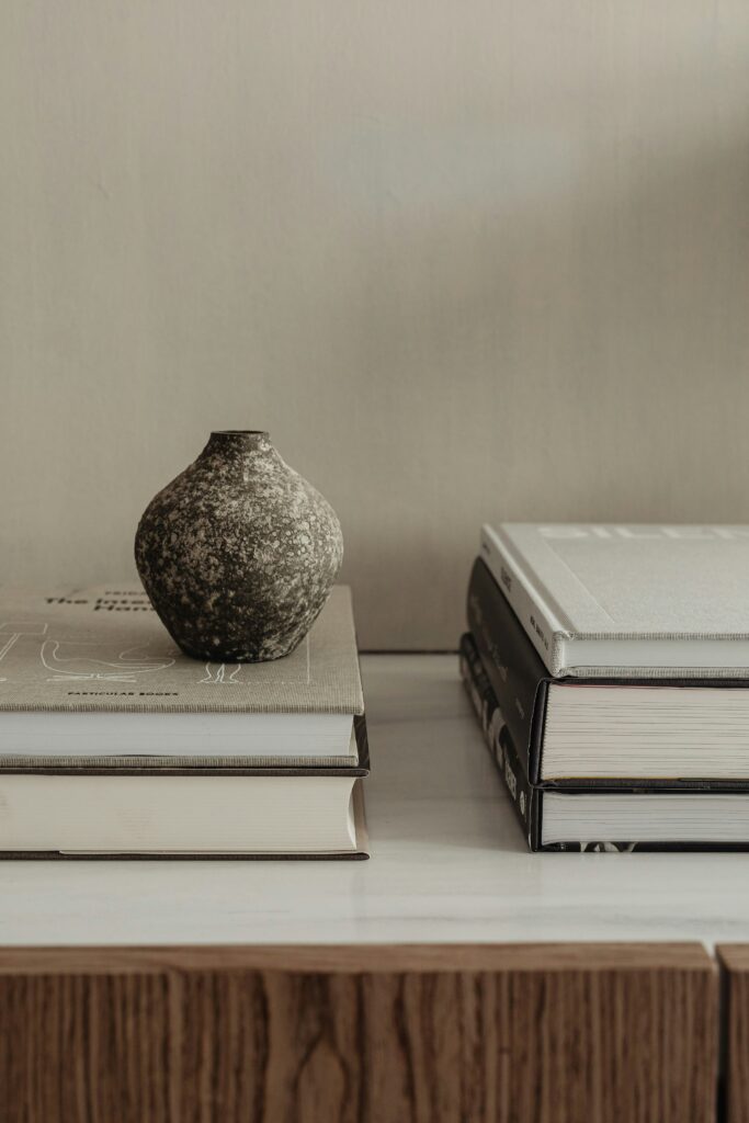 A ceramic vase resting on stacked neutral-toned books beside a small pile of hardcover volumes on a minimalist wooden shelf.